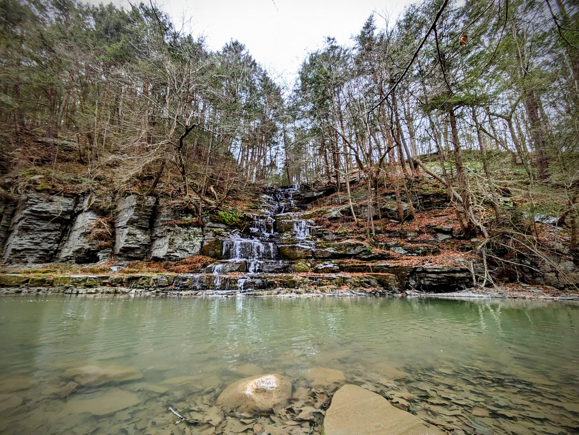 A tiered waterfall spilling over shale ledges into a clear green plunge pool, in a wooded Finger Lakes gorge